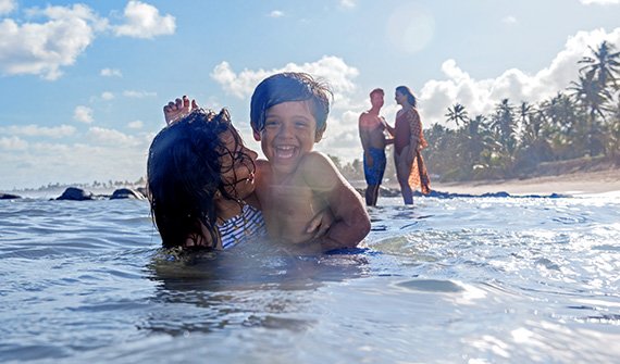 Família se divertindo na praia em frente ao Costa do Sauípe, com crianças brincando na areia e pais relaxando ao fundo