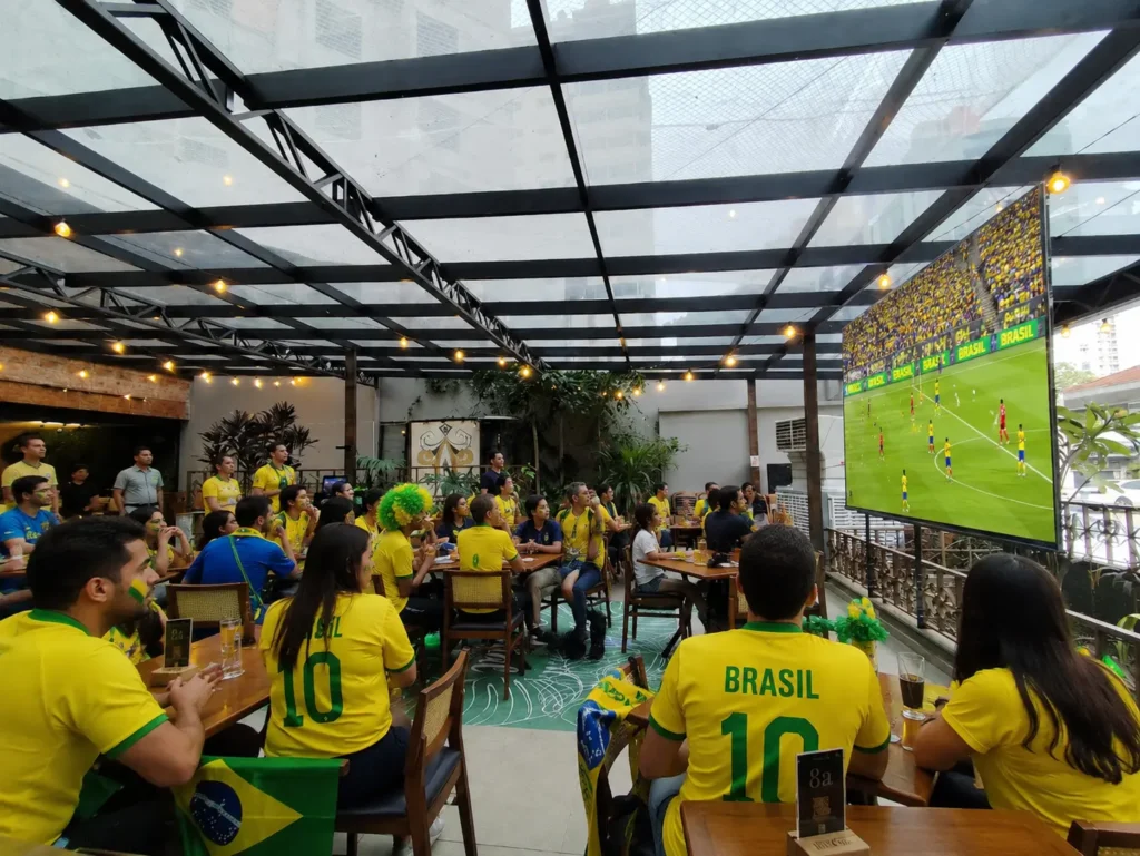 Família assistindo a um jogo de futebol em ambiente festivo no Quintal Del Carmem em Santo André, com telões de LED e decoração temática da Copa.