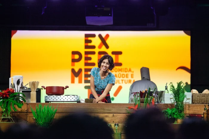 Bela Gil em evento de gastronomia no SESC, rodeada por alimentos frescos e saudáveis.